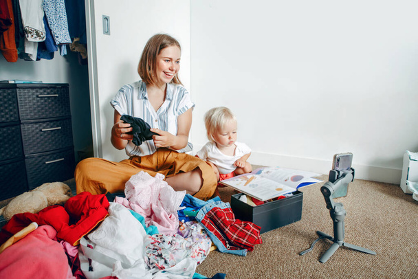 Young mother blogger doing video live stream for social media while sorting clothes at home. Video call chat with friends and family on smartphone. Woman using gimbal tripod for video and phone calls. - Photo, Image