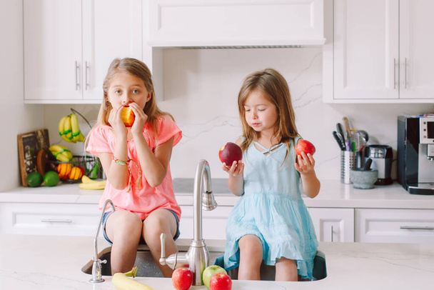 Caucasian children girls eating fresh fruits sitting in kitchen sink. Happy family sisters siblings having snack. Organic food and healthy delicious tasty meal for kids. Lifestyle authentic moment. - Photo, Image