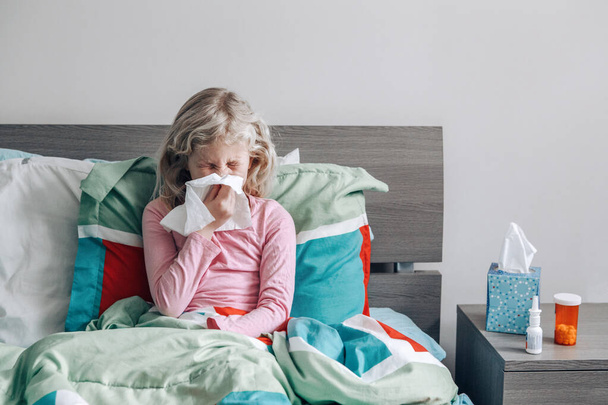 Niña caucásica enferma con fiebre acostada en la cama sonándose la nariz con papel de servilleta. Enfermedad gripal por virus. Concepto de medicina y salud - Foto, Imagen
