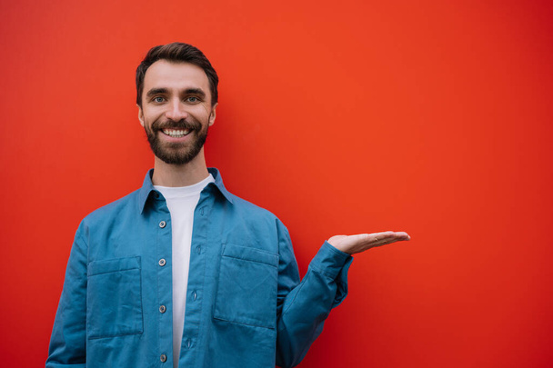 Portrait of young smiling man wearing casual shirt pointing with hand, isolated on red background. Copy space, advertising concept  - Valokuva, kuva