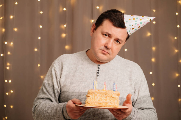 sad man in festive cap with birthday cake blows out candles.sadness due to aging - Photo, image