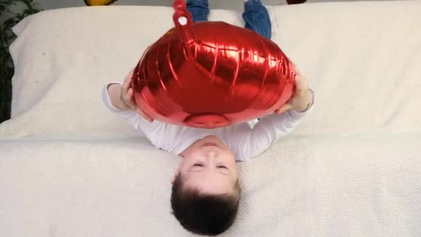 Um menino bonito no sofá está jogando de cabeça para baixo e se divertindo com uma bola em forma de coração vermelho. Feriado Dia dos Namorados. Movimento lento - Filmagem, Vídeo