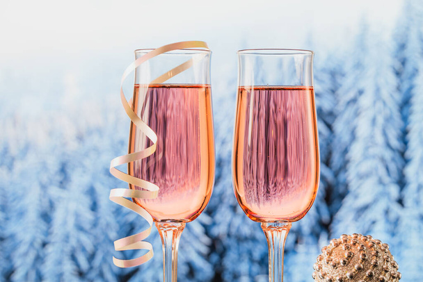 Two glasses of rose champagne and Christmas or New Year decoration with winter snowy forest bokeh as a background. Romantic dinner. Winter holiday concept. - Foto, imagen