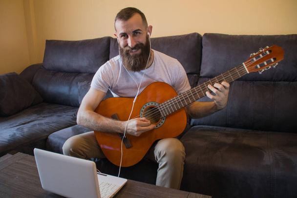 Sonriente hombre barbudo aprendiendo a tocar la guitarra con auriculares y portátil - Foto, imagen