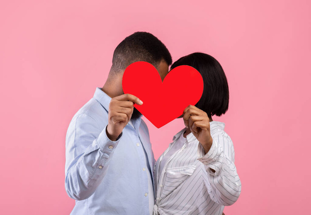 Novios románticos escondidos detrás del corazón de papel rojo y besándose en el fondo del estudio rosa - Foto, Imagen