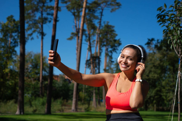 Atractiva mujer en forma con auriculares y teléfono móvil en la mano sonríe a la cámara mientras se relaja en el fondo del bosque de pinos - Foto, Imagen