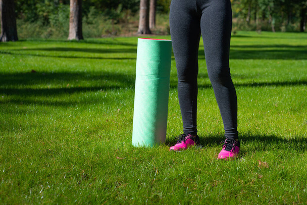 Περικοπή θέα στα πόδια της γυναίκας στέκεται κοντά σε ένα στρώμα γιόγκα σε ένα πράσινο γρασίδι στο πάρκο σε μια ηλιόλουστη μέρα. Έννοια της γυμναστικής και γιόγκα - Φωτογραφία, εικόνα