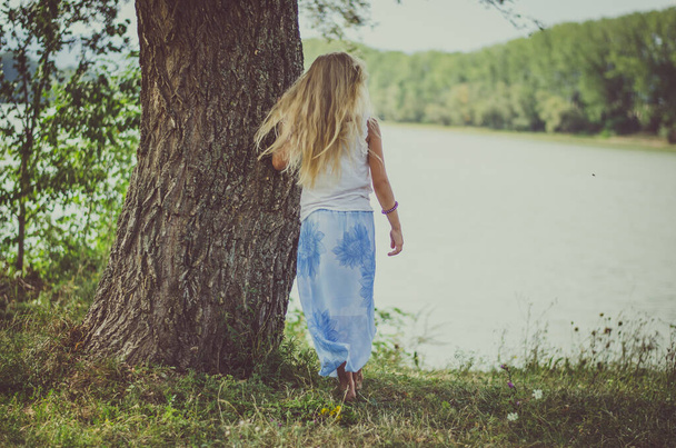 池のそばの木のそばに長いブロンドの髪の子供が立っていて - 写真・画像