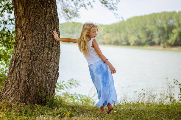 αξιολάτρευτο παιδί με μακριά ξανθά μαλλιά στέκεται δίπλα στο δέντρο δίπλα στη λίμνη - Φωτογραφία, εικόνα