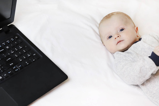 A cute baby is lying on the bed next to a laptop. Parents sit at the computer and look after the child. Copy space - technology concept, home office, remote work, millennial with son, busy man - Photo, Image
