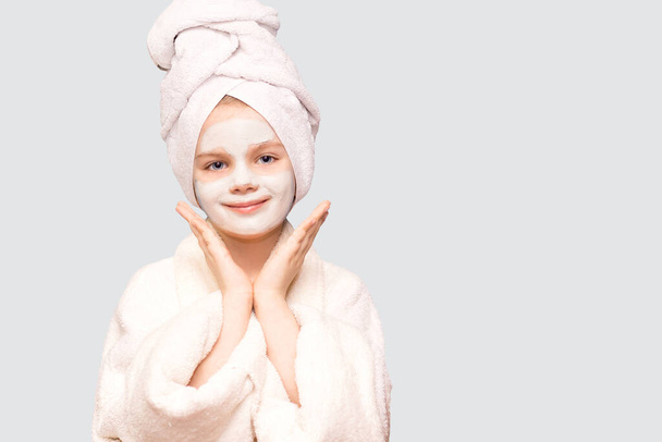 Fille enfant agréable avec serviette enveloppée sur la tête expression ravie isolé sur blanc, applique un produit de beauté naturel sur le visage, débouche les pores, la peau saine - Photo, image