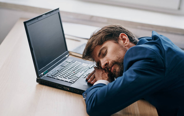 Man in een pak aan een werktafel voor een laptop kantoor moe - Foto, afbeelding