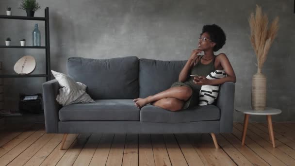 Heureuse jeune femme afro-américaine s'asseoir se détendre sur un canapé confortable et heureux avec le message au téléphone - Séquence, vidéo