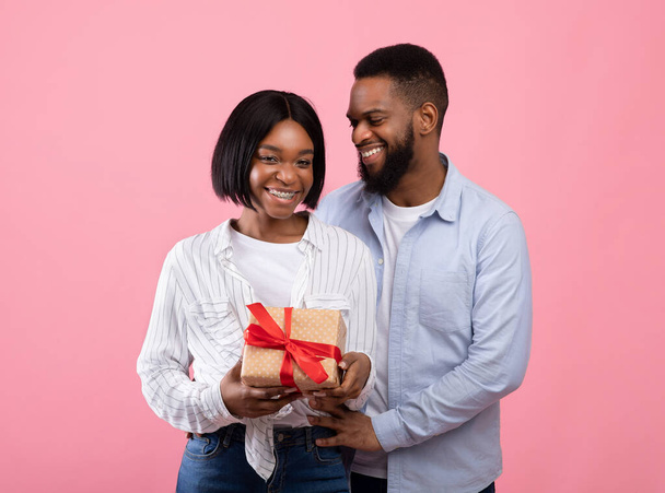 Loving black couple holding wrapped gift for Valentines Day, congratulating each other with holiday on pink background - Φωτογραφία, εικόνα