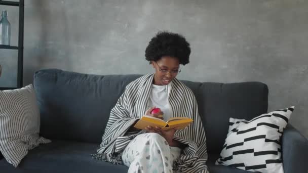 Fröhliche afroamerikanische junge Frau sitzt entspannt auf der gemütlichen Couch und liest ein Buch. Froh über Umzug in neue Wohnung. - Filmmaterial, Video