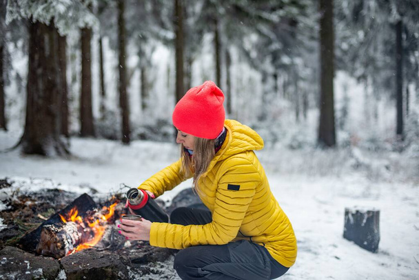 Um turista loiro em uma jaqueta amarela derrama chá quente de uma garrafa térmica em uma caneca de metal perto do fogo. - Foto, Imagem