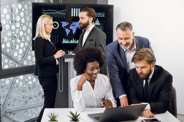 Positive purposeful multiracial business colleagues, working together over joint business project. Focus on mature blond woman and bearded man behind, using interactive screen in modern meeting room - Foto, Bild