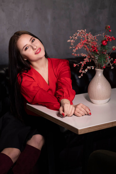 赤いブラウスと装飾的な輝く赤い枝を持つ花瓶の近くのロフトヴィンテージカフェで黒いスカートのセクシーな魅力的なブルネットの女性。コピースペース。バレンタインデー  - 写真・画像