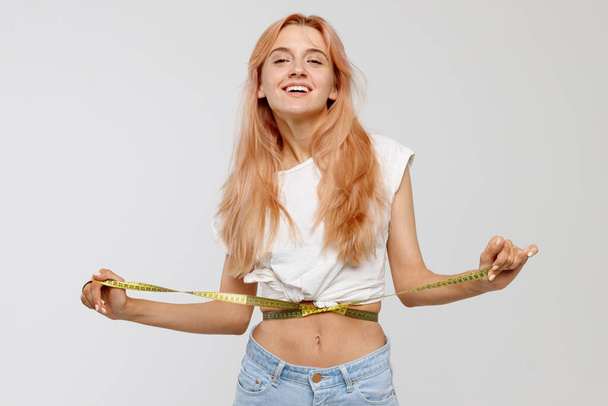 Стройная счастливая молодая женщина с жёлтой рулеткой, измеряющая свою тонкую талию, смотрящая в камеру, изолированная на сером фоне. Потеря веса, диета, контур тела - Фото, изображение