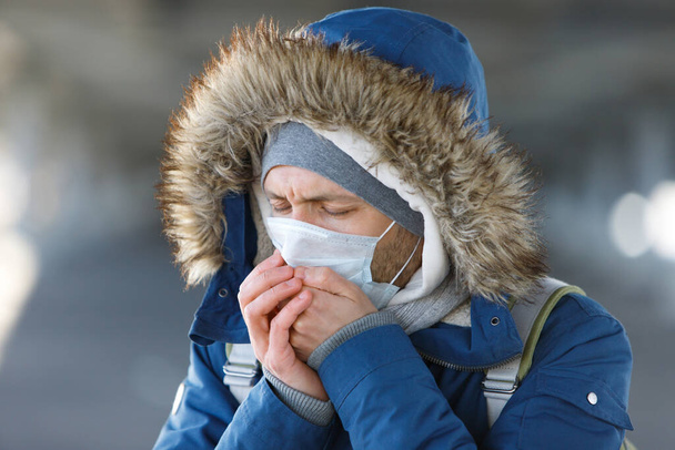 冷たい、くしゃみをし、咳をし、屋外で医療面マスクを身に着けている病気の若い男のクローズアップ。気管支炎、病気、インフルエンザの流行 - 写真・画像