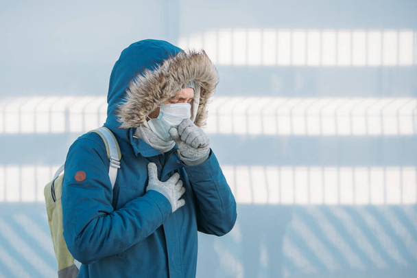 Portrait of sick young man in blue jacket put on a hood, having a cold, feeling unwell, coughing, wearing medical face mask, outdoors. Illness, upcoming flu season.  - Photo, Image