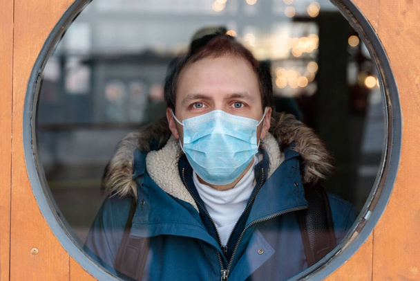 L'uomo guarda fuori dalla finestra rotonda, indossa una maschera medica protettiva contro le malattie infettive trasmissibili e come protezione contro l'influenza o il coronavirus, covid-19 in luogo pubblico. Concetto di quarantena.  - Foto, immagini