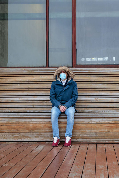 Больной человек в капюшоне сидит один на скамейке, в защитной маске от инфекционных заболеваний, ковид-19. Пустой город от людей и туристов из-за пандемии коронавируса - Фото, изображение