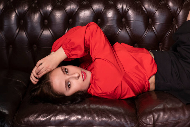 retrato de mujer morena atractiva en blusa roja y falda negra acostada en sofá de cuero en un café vintage. San Valentín. espacio de copia. - Foto, imagen