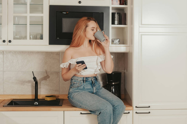 Kobieta siedzi w domu w kuchni. Dziewczyna z dobrą figurą pije herbatę i patrzy na telefon. Dostawa żywności, sklepy mobilne, zakupy online. Pije herbatę lub kawę - Zdjęcie, obraz