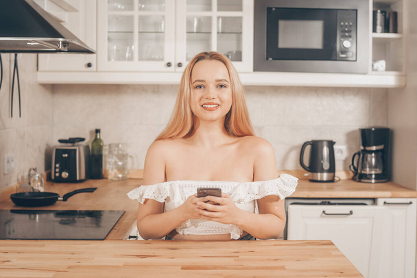Piękna kobieta siedzi przy stole na tle kuchni w domu. Dziewczyna ze smartfonem w rękach. Blondynka pisze wiadomość albo zamawia na wynos. Dostawa online - Zdjęcie, obraz