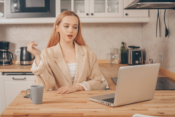 Woman behind laptop works from home. A beautiful blonde in a jacket speaks by video call. The student submits a report online. The girl teaches courses, lectures on the Internet. Freelance earnings. The model is gesturing. - Photo, Image
