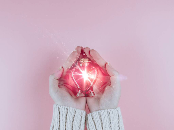 A woman holds in her palms a bottle in the shape of a heart with a red drink in the middle on a pink background, top view close-up. - Foto, Imagen