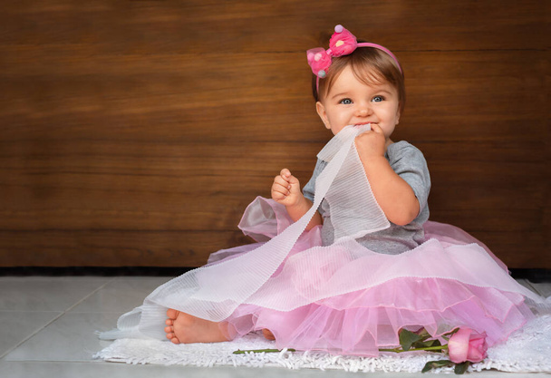 Baby in een jurk en linten op een houten achtergrond. Een kind in een roze jurk bijt een wit lint op een houten achtergrond - Foto, afbeelding