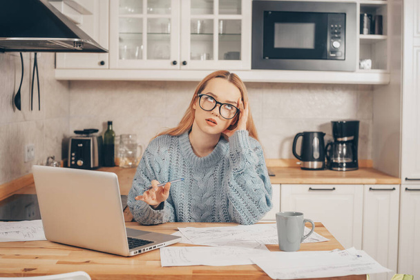 A woman works from home using a laptop. A beautiful blonde in a sweater and glasses, there is a mug and papers on the table. Home schooling, work online. Displeased, tired, indignant student. - Foto, Imagen