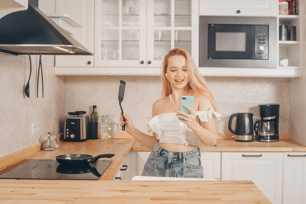 A girl in headphones with a phone stands in the kitchen and prepares food behind an induction stove. Woman singing and dancing in the kitchen with a smartphone, music. - Photo, Image