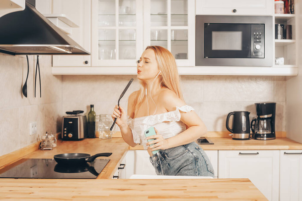  Γυναίκα τραγουδούν και χορεύουν στην κουζίνα με ένα smartphone, μουσική.Ένα κορίτσι με ακουστικά με ένα τηλέφωνο στέκεται στην κουζίνα και ετοιμάζει το φαγητό πίσω από μια επαγωγική σόμπα. - Φωτογραφία, εικόνα