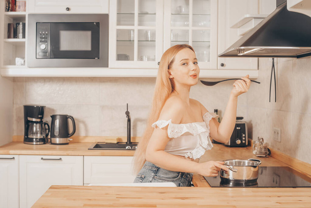 Beautiful girl prepares food on an induction stove. A woman cooks soup or sauce in a saucepan. The blonde in the bright kitchen tastes. - Valokuva, kuva