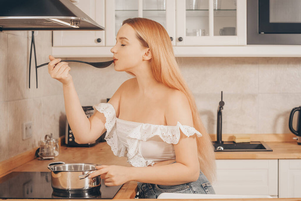 Mooi meisje bereidt eten op een inductiekachel. Een vrouw kookt soep of saus in een pan. De blonde in de lichte keuken smaakt. - Foto, afbeelding