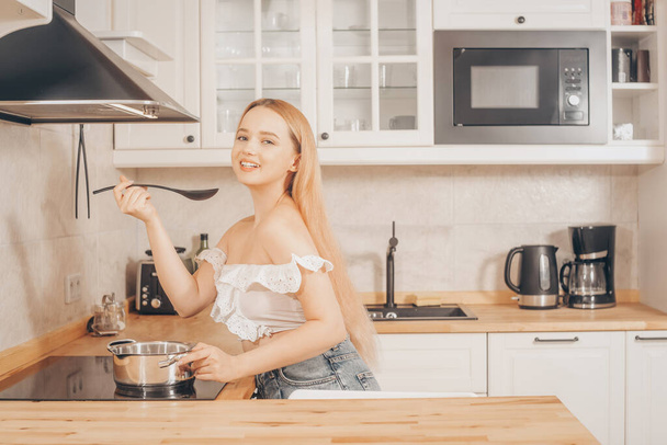 Beautiful girl prepares food on an induction stove. A woman cooks soup or sauce in a saucepan. The blonde in the bright kitchen tastes. - Photo, Image