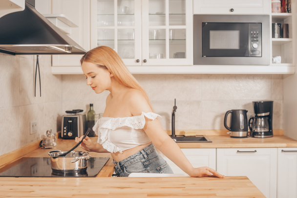 Beautiful girl prepares food on an induction stove. A woman cooks soup or sauce in a saucepan. The blonde in the bright kitchen. - Photo, Image