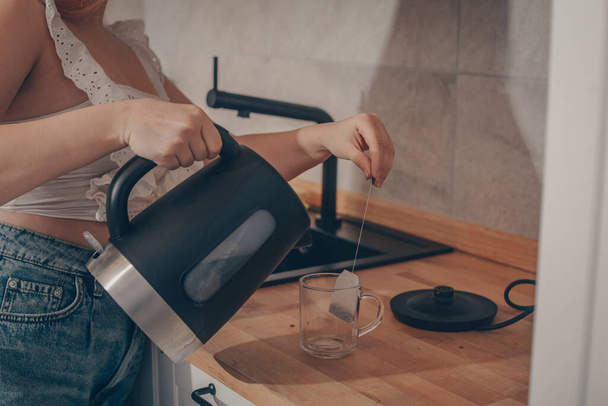 Hay un hervidor negro en la mesa de la cocina. La mano de una mujer sostiene una tetera y hace té. Bolsa de té en una taza. - Foto, Imagen