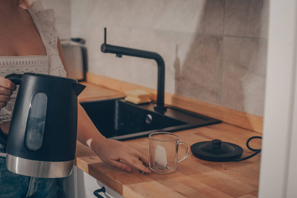 Auf dem Küchentisch steht ein schwarzer Wasserkocher. Die Hand einer Frau hält eine Teekanne und kocht Tee. Teebeutel im Becher. - Foto, Bild