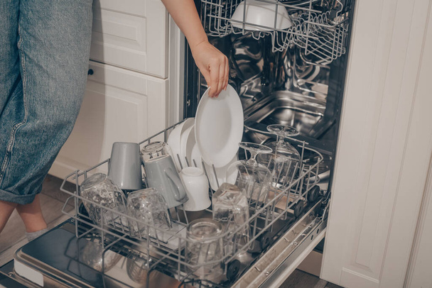 Built-in dishwasher, dishwashing. A woman loads washed dishes, cups, glasses. A woman's gentle hand puts something in the dishwasher or pulls out, unloads. - Foto, Bild