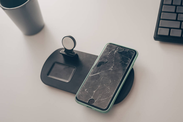 Το κινητό τηλέφωνο είναι στο τραπέζι. Smartphone με σπασμένη οθόνη, γυαλί. Επισκευή οθόνης, αντικατάσταση γυαλιού, επιδιόρθωση. Τηλέφωνο για ασύρματη φόρτιση - Φωτογραφία, εικόνα