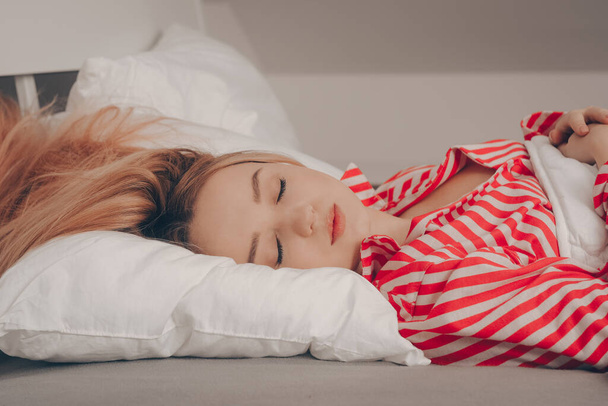 Śpi piękna dziewczyna na białym łóżku w czerwonej piżamie w paski. Zdrowy sen. Kobieta z zamkniętymi oczami i długimi sutr włosami na łóżku. - Zdjęcie, obraz