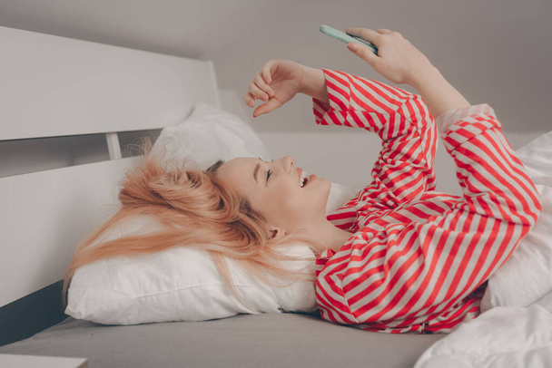 Een meisje kijkt 's morgens naar de telefoon, een vrouw in bed kijkt naar een video of leest het nieuws. Internetverslaving. Het meisje kijkt naar de inhoud en lacht. - Foto, afbeelding