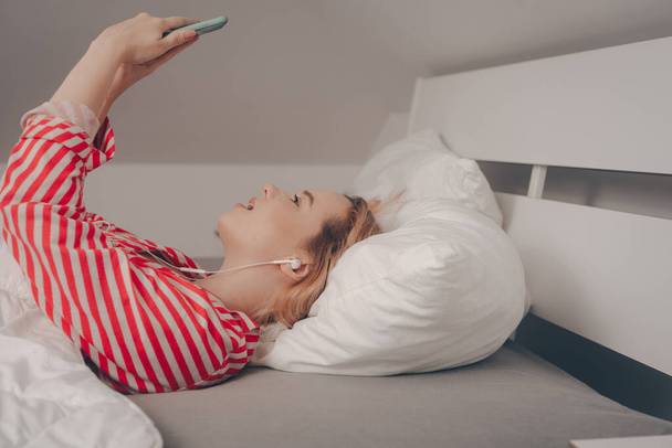 Девушка ложится на кровать перед сном. Женщина в наушниках смотрит в телефон, говорит по видеосвязи, снимает видео. - Фото, изображение