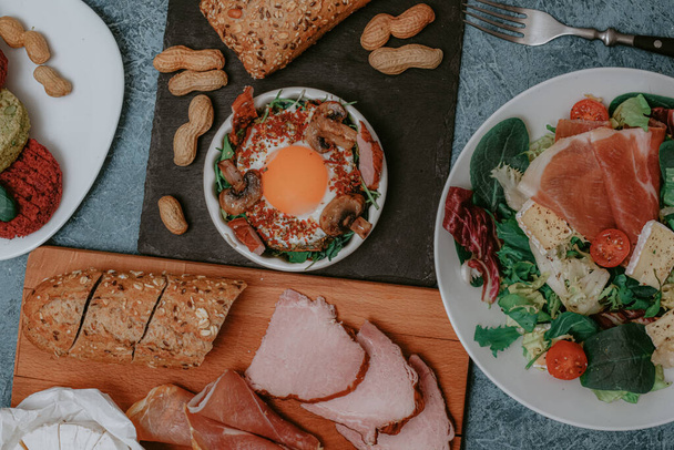 Lots of food and dishes in the background. Meat platter and Camembert cheese with baguette. Salad with prosciutto. Perfect fried eggs with mushrooms and spices. Vegan vegetable cutlets and peanuts. Fork. Food delivery. - Photo, Image