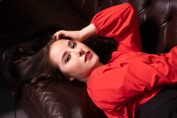 muotokuva houkutteleva brunette nainen punainen pusero makaa nahkasohvalla. Ystävänpäiväkortteja. kopioi tila. - Valokuva, kuva