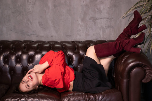 retrato de mulher morena atraente em blusa vermelha e saia preta deitada no sofá de couro em um café vintage. São Valentim. espaço de cópia. - Foto, Imagem